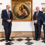 Three men standing beside a painting of a woman holding a baby.
