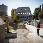 A cylindrical building, partially destroyed, is seen in the distance of a color photograph. It is perhaps five stories tall. In the foreground, two people in orange vests walk through a construction site.