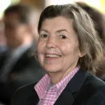 A smiling woman in a pink crosshatched shirt wearing a suit jacket.