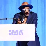 A man in a blue suit and a hat speaks into a microphone at a podium with 