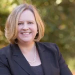 A smiling blonde white woman wearing a string of pearls and a blazer.