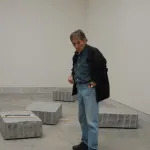 A white man in jeans, a button down shirt, and a black jacket standing amid a room filled with rough-hewn stones on its floor.