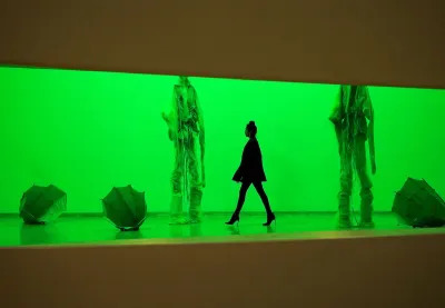 A woman walking through a gallery lit a bright shade of green. She walks beneath two sculptures of towering figures draped in fabric. Around them are objects resembling inverted umbrellas.
