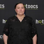 An Indigenous woman stands on a red carpet before a backdrop that reads 