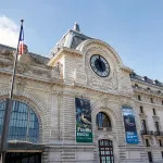 A grand Beaux-Arts building of around six stories is seen on a clear day in a color photograph. There is a large clock at the center of its soaring facade.