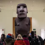 A large basalt sculpture of a shirtless figure with a large face. The sculpture towers above people who stand below.