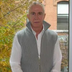 A man in a grey vest seated before a window looking out onto a tree.