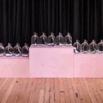 Three award-ceremony-like platforms in pink hold five glass jugs filled with a white ribbon.