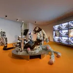 An installation composed of stuffed replicas of appendages on a platform facing TV screens.