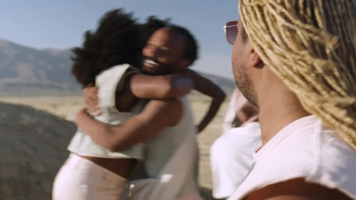 Two Black people in white clothing hug in front of a desert landscape, while another person looks on.