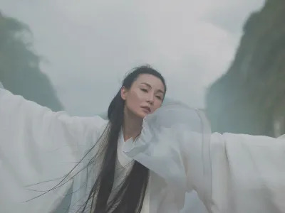 A woman in flowy fabric waving her arms in a mountainous landscape.