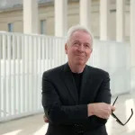 An older man in a black suit jacket and black shirt looks up with a slight smile as he holds black sunglasses in his right hand, with arms crossed. White architecture—many thin columns and a kind of fence stand behind him.