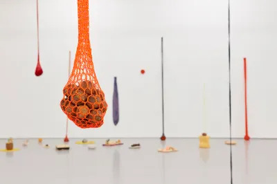 A sculpture resembling a pile of rocks suspended in hanging orange netting. Other similar works hang behind it.
