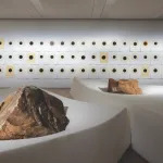 In a large gallery, two boulders rest on top of thick rectangles of white foam. Along the back wall are three rows of works on paper, all including a black circle with a yellow outer circle.