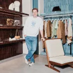 Portrait of Jonathan Anderson in a Loewe retail store. He wears a crewneck sweater and light-wash jeans and leans on a wood chair.
