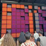 People gaze at a 25-foot-tall installation made of woven squares in red, blue, and gold.