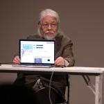An elderly Asian man holding up a laptop screen to an audience.
