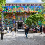 People walking in and out of a treed courtyard in front of a brightly painted building with columns. On its roof, there is the phrase 