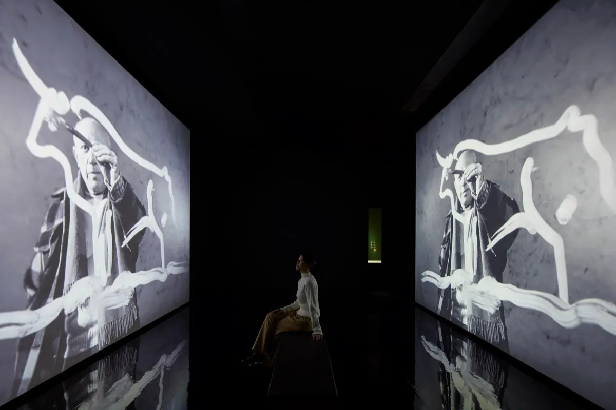A woman watches a screen depicting a man painting.