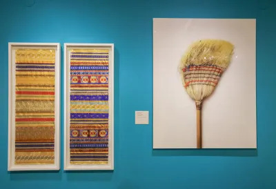 View of a museum exhibition with a photograph of a broom at right. At left two tapestries made of found beer labels. 