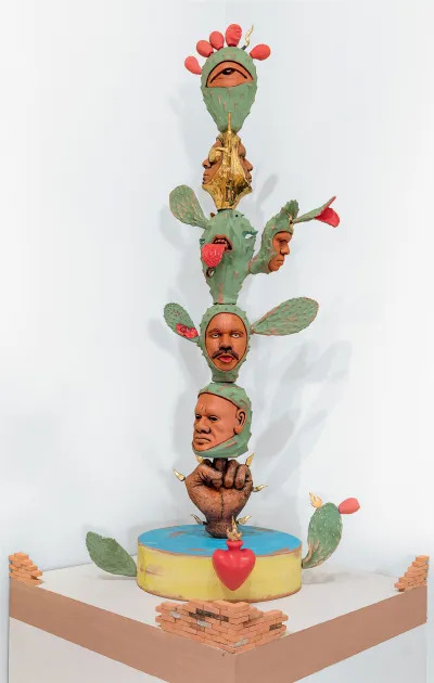 A sculpture in with a fist holds up a cactus/nopal in which faces, an eye, and a mouth emerge. 