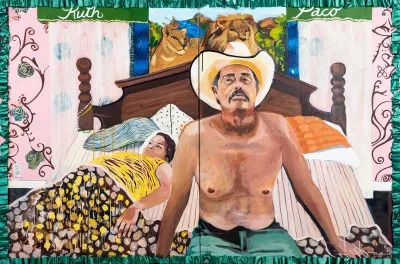A shirtless man wearing a cowboy hat sits on a bed, with a woman behind him under the cover. 