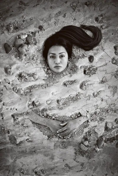 A black-and-white photograph of an Indigenous woman buried in cracked sand. Her face and hands are unburied. She stares at the viewer.