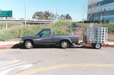 A 1994 Toyota Pickup towing a sculpture that measures air quality. A green freeway sign is seen in the background.
