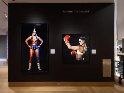 View of a museum wall that is painted black. On it hang two color photographs of Indigenous women, one dressed as Wonder Woman and the other as a boxer.