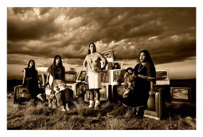 A sepia photograph showing four Indigenous adults and a baby in a landscape. behind them are a collection of TVs showing stereotyped media of Indigenous people.