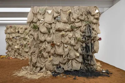 Two towers features rows of burlaps sacks that hold various objects, including dried flowers and feathers. One area is smeared with a black, ashy substance. The towers are planted into a ground covered in dirt.
