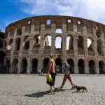 Two women walk a dog past an ancient building.