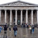 A classical looking structure with columns and people walking in front of it.