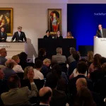 A scene from an auction: a man stands at a rostrum next to several people and in front of a crowd.