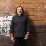 A man pictured against a graffitied brick wall.