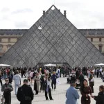 A crowd of people before a glass pyramid.