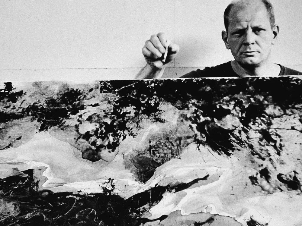 American abstract expressionist painter Jackson Pollock (1912 - 1956), holding a cigarette, stands behind one of his paintings in his studio at