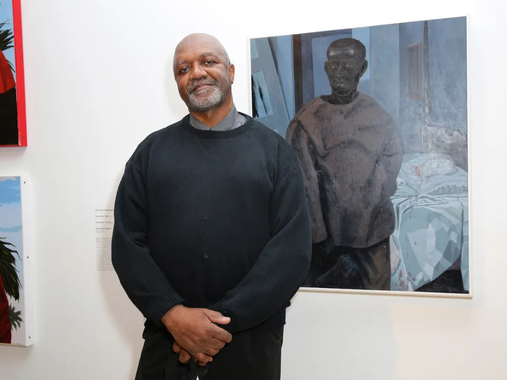 A Black man in a black sweater standing beside a painting of a Black man in similar clothes.