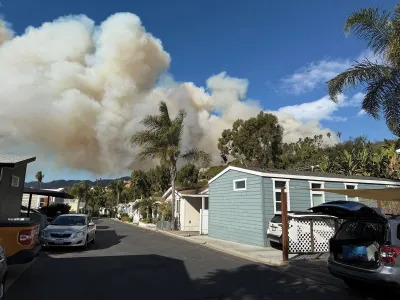 A residential street on a sunny with the smoke of an approaching wildfire visible in the center background. 