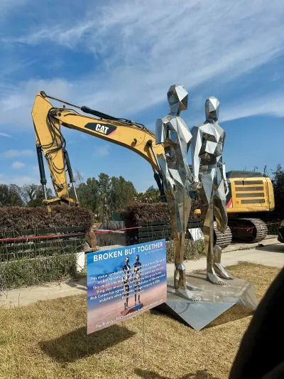 A reflective metal sculpture made of various polygons that resemble two standing figures holding hands. A crane sifting through a burned home is seen in the background. 