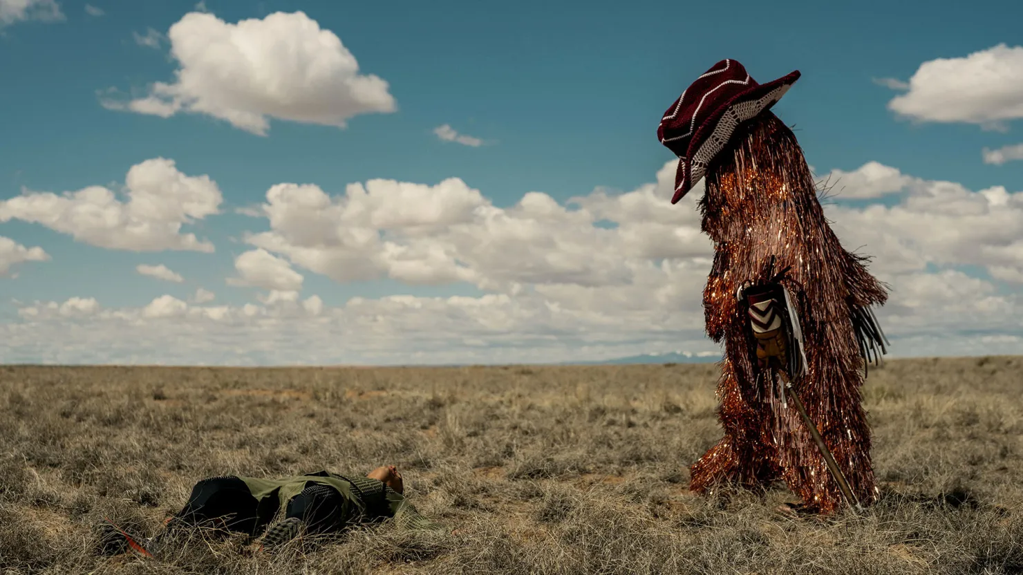A mascot covered in tinsel and wearing an oversize cowboy hat walks across a desert.